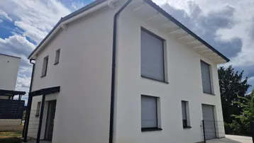 Modernes Einfamilienhaus mit weißer Fassade, dunklem Dach und Rollläden an den Fenstern.