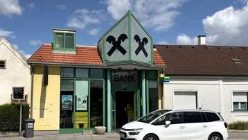 Außenansicht eines Bankgebäudes mit markantem Giebel und großen Glasflächen, ein Auto parkt davor.