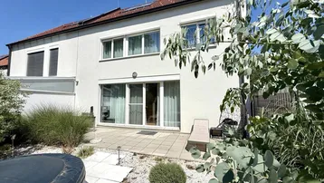 Hausfassade mit großer Terrasse, Whirlpool und Gartenbereich unter blauem Himmel.