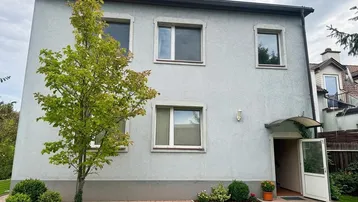Vorderansicht des Hauses mit hellgrauer Fassade, Fenstern und Eingangsbereich.