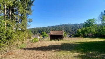Weitläufiges Grundstück mit kleinem Schuppen und Blick auf bewaldete Hügel unter blauem Himmel.