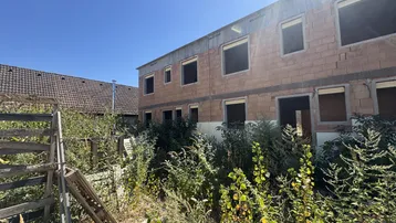 Unfertiges Gebäude mit Ziegelwänden und leeren Fensteröffnungen auf einer Baustelle.