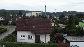 Luftaufnahme einer Wohnstraße mit Einfamilienhaus, viel Grün und Industrieanlagen im Hintergrund.