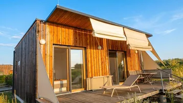 Modernes Holzhaus mit großer Holzterrasse, Sonnenliegen und Beschattung unter blauem Himmel.