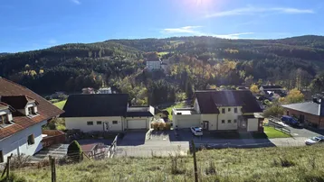 Panoramablick auf Wohnhäuser in einer ländlichen Umgebung mit bewaldeten Hügeln und strahlendem Sonnenschein.
