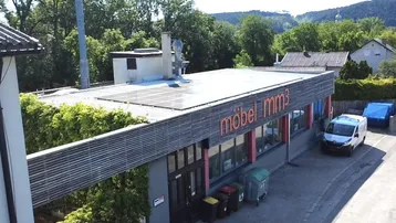Außenansicht eines Geschäftsgebäudes mit Solarpaneelen auf dem Dach und einem Lieferwagen davor. Umgeben von Bäumen und Hügeln.
