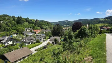 Panoramablick über eine grüne Wohnsiedlung in einer hügeligen Landschaft unter blauem Himmel.