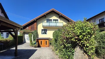 Frontansicht des Hauses mit gepflasterter Auffahrt, Balkon und üppiger Bepflanzung.