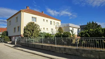 Großes Wohngebäude mit heller Fassade und umzäuntem Vorgarten an einer ruhigen Straße.