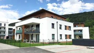 Modernes Mehrfamilienhaus mit weißen Fassaden, Balkonen und Blick auf die grüne Berglandschaft.
