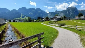 Idyllischer Feldweg entlang eines Baches mit grünen Wiesen und majestätischen Bergen im Hintergrund.