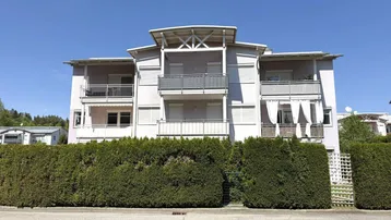 Mehrfamilienhaus mit mehreren Balkonen und gepflegter Hecke im Vordergrund unter blauem Himmel.
