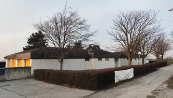 Gebäudeansicht mit flachem Dach, hellen Wänden und kahlen Bäumen, umgeben von einer Hecke.