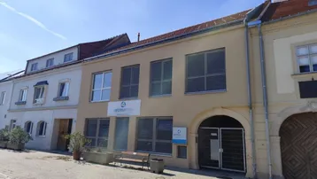 Gebäude mit beiger Fassade, blauen Fensterläden und einem Torbogen am Eingangsbereich, umgeben von weiteren Häusern.