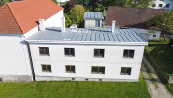 Luftaufnahme eines weißen Gebäudes mit grauem Blechdach und umliegender Grünfläche.