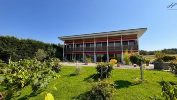 Modernes Gebäude mit großen Fensterfronten, umgeben von einem gepflegten Garten und Weinreben.