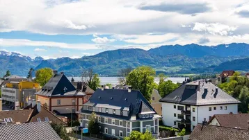 Panoramablick über eine Stadt mit See und Bergen im Hintergrund unter blauem Himmel.