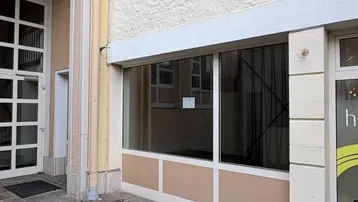 Ansicht der Gebäudefassade mit großen Fenstern und Eingangsbereich, gepflasterter Gehweg davor.