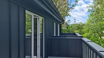 Moderner Balkon mit dunkler Verkleidung, hellen Fliesen und Blick ins Grüne sowie auf ein Gebäude.