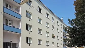 Fassade eines Mehrfamilienhauses mit hellen Wänden und blauen Balkonen an einem sonnigen Tag.
