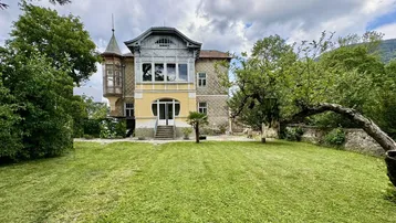 Historische Villa mit großem Garten und altem Baumbestand unter bewölktem Himmel.