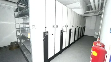 Lagerraum mit mobilen Regalsystemen und sichtbaren Rohrleitungen an der Decke, ausgestattet mit Feuerlöschern.