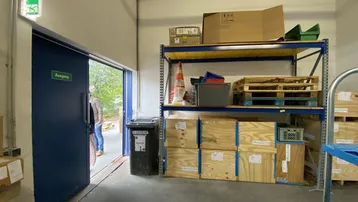Lagerbereich mit Regalen, Holzkisten und offenem Ausgang ins Freie, gut beleuchtet durch die Tür.