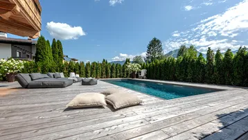 Großer Außenpool mit Holzdeck und bequemen Lounge-Möbeln, umgeben von einer grünen Hecke und Bergblick.
