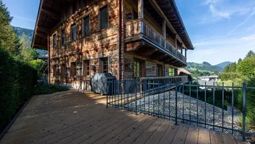 Rustikales Holzhaus mit großer Holzterrasse und Bergblick, umgeben von grüner Natur.
