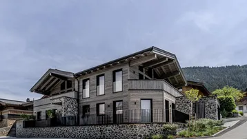 Modernes Chalet mit Holz- und Steinfassade, großen Fenstern und Balkonen, eingebettet in eine Berglandschaft.