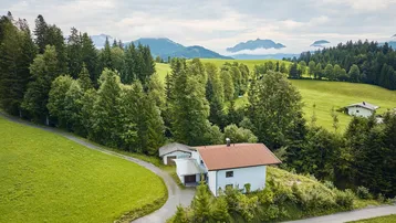 Luftaufnahme eines Hauses in einer idyllischen grünen Hügellandschaft mit Bergen im Hintergrund.