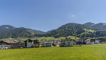 Weitläufiger Blick über eine grüne Wiese auf ein charmantes Dorf und die umliegenden Berge.