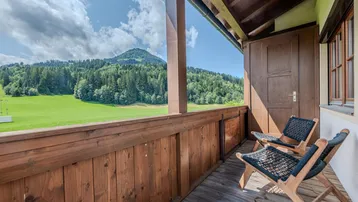 Sonniger Holzbalkon mit bequemen Stühlen und weitem Blick auf die grüne Berglandschaft.