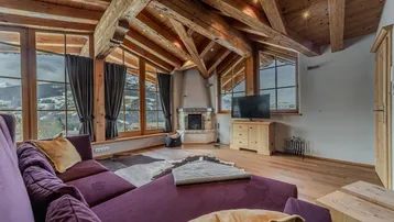 Rustikales Wohnzimmer mit Kamin, Holzbalkendecke und großen Fenstern mit Blick auf die verschneite Landschaft.