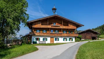 Traditionelles bayerisches Bauernhaus mit Holzbalkonen und grünen Fensterläden, umgeben von grüner Landschaft.