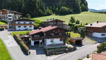Traditionelles Haus mit Holzbalkonen und rotem Ziegeldach in ländlicher Umgebung mit viel Grün.