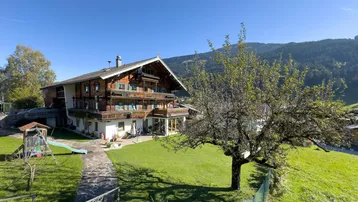 Traditionelles Holzhaus im alpinen Stil mit großem Garten, Spielplatz und Bergblick unter blauem Himmel.