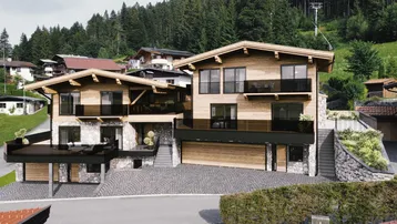Moderne Chalet-Architektur mit Holz- und Steinfassade, umgeben von grünen Bergen und Wäldern.