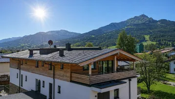 Modernes Gebäude mit Holzfassade und Balkonen vor einer beeindruckenden Bergkulisse unter strahlendem Sonnenschein.