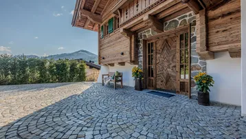 Traditionelles Holzhaus mit Steinfassade und gepflastertem Eingangsbereich, umgeben von grüner Natur.