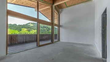 Heller, unmöblierter Innenraum mit großen Fenstern und Holzbalkendecke, bietet einen weiten Blick auf die grüne Berglandschaft.