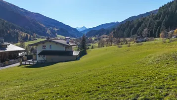 Haus in einer malerischen Berglandschaft mit weiten grünen Wiesen und bewaldeten Hängen unter blauem Himmel.