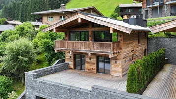 Modernes Chalet mit Holzfassade, großzügiger Terrasse und Balkon, umgeben von grüner Natur und Bergen.