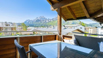 Sonniger Balkon mit Rattanmöbeln, Glastisch und herrlichem Bergblick über die Dächer der Stadt.