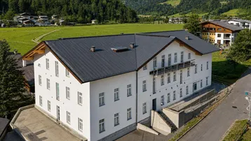 Modernes Gebäude mit weißer Fassade und dunklem Satteldach, umgeben von grünen Wiesen und Bergen.