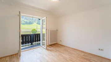 Helles Zimmer mit Holzboden und Zugang zu einem Balkon mit Blick ins Grüne.