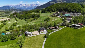 Luftaufnahme einer Wohngegend mit Golfplatz und Alpenpanorama im Hintergrund unter blauem Himmel.