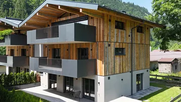 Modernes Mehrfamilienhaus mit Holzfassade, Balkonen und Bergblick in grüner Umgebung.