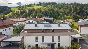 Luftaufnahme eines modernen Mehrfamilienhauses in alpiner Umgebung mit umliegenden Bergen.
