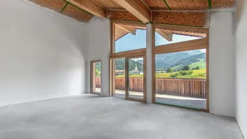 Heller, unmöblierter Wohnraum mit hohen Decken, Holzbalken und großen Fenstern mit Bergblick.